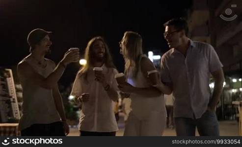 Slow motion steadicam shot of happy friends of three men and a woman toasting with take-away coffee cups. They are laughing, drinking coffee, and walking in the night city street