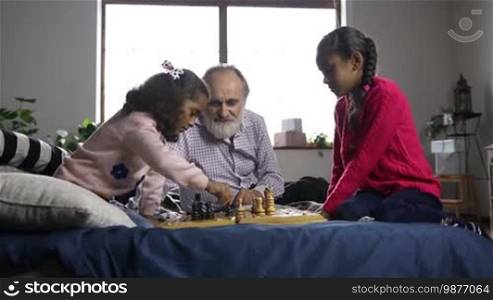 Smart little mixed race girl captured a pawn and celebrating with joyful grandfather, giving high five while playing chess with her sister at home. Two sweet girls playing chess game with their grandpa in domestic interior. Dolly shot. Slow motion.
