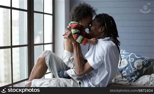 Smiling African American daddy playing with his little toddler boy in the bedroom at home. Joyful African man with dreadlocks holding cute mixed race son and having fun on the bed.