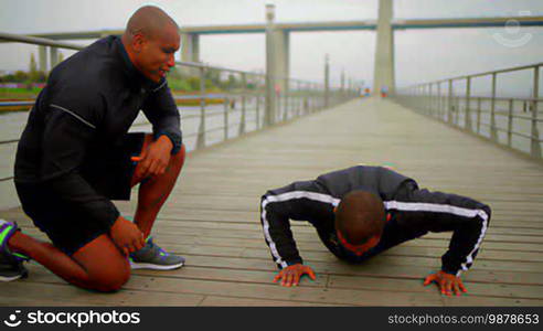 Smiling bald man motivating his friend who is doing push-ups. Two people training on a wooden pier. Sport and supporting concept