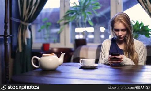 Smiling blonde teenage female texting on cell phone while sitting in modern coffee shop and drinking tea. Beautiful young hipster woman using mobile phone while enjoying her free time in expensive stylish cafe.