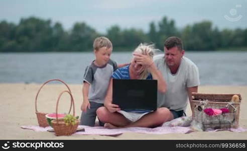 Smiling family talking with grandparents using laptop during summer holidays on the beach. Cheerful family of three chatting and sending kisses to grandparents using notebook via skype