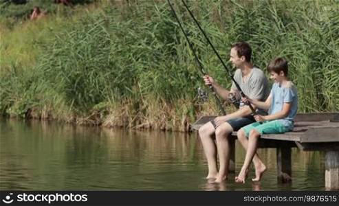 Smiling father and teenage boy sitting on wooden pier swung with their legs in water while fishing together by the lake in summer day. Positive dad communicating with son while fishing with rods near pond over beautiful landscape background. Slow motion.