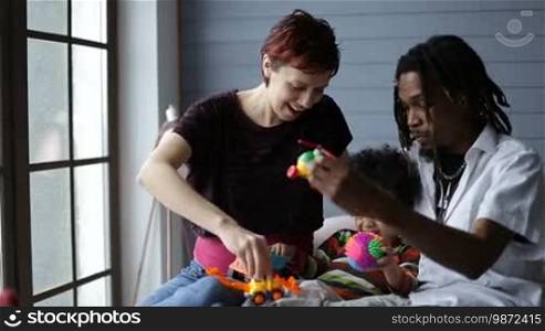 Smiling interracial family with sweet little toddler son enjoying time together at domestic interior. Multiethnic parents with mixed race adorable child playing with colorful toys while sitting on the bed in bedroom.