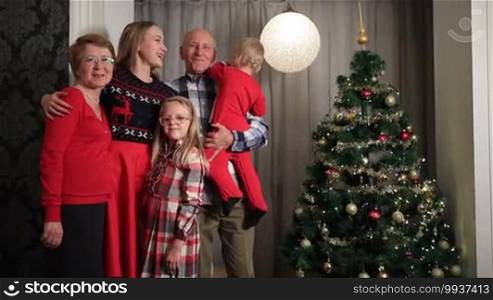 Smiling multi-generation family standing by Christmas tree at home. Happy smiling family with grandparents and children at home celebrating New Year and enjoying winter holiday together.