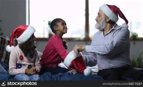 Smiling small mixed race girls reaching into Santa hat held by joyful grandfather and picking up pieces of paper with player names during happy family playing Secret Santa on Christmas Eve. Joyful family spending leisure on winter holidays.