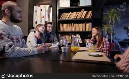 Smiling teenagers sitting at cafe table and enjoying great time together. Group of young joyful friends drinking coffee and juice while hanging out in coffee shop. Hipsters spending leisure time in cafeteria, chatting and laughing.