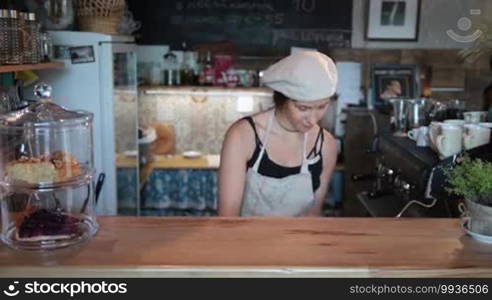 Smiling waitress in apron putting on counter a tasty cheesecake on cake stand. Attractive female barista opening a cake stand with delicious cheesecake and giving thumb up