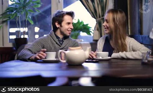 Smiling young couple in love sitting at cafe table and drinking hot cup of tea while hanging out and talking at the restaurant. Two lovers enjoying time together and drinking tea in cozy coffee shop while communicating with each other.