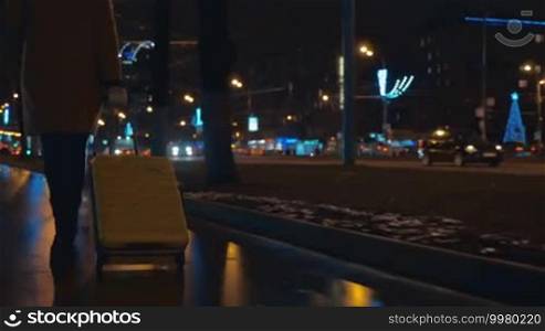 Steadicam back and low angle shot of a woman walking in the evening city with trolley bag on background of luminous city and passing cars