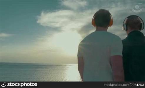 Steadicam back shot of young couple in wireless earphones looking at scenic view with blue sea and bright evening sun reflecting in water