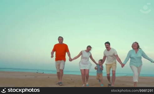 Steadicam shot of a big family of parents, son, and grandparents holding hands and running on the beach. Happy vacation together