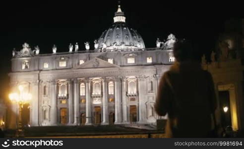 Steadicam shot of a female tourist with a pad near St. Peter's Basilica in Vatican City at night. She is making a video or taking photos of the greatest church of Christendom