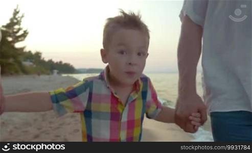 Steadicam shot of a happy little boy in a colorful checked shirt holding grandparents' hands while they walk along the seaside, view of a barefoot family walking on the sand