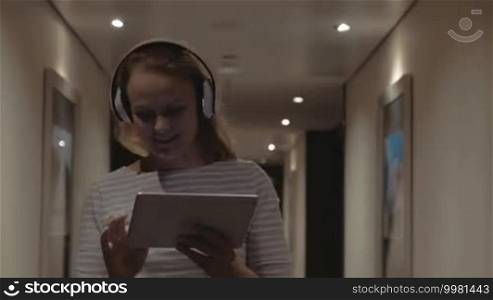 Steadicam shot of a young happy woman walking down hotel corridor with tablet PC and enjoying music in wireless headphones. Cheerful girl dancing and singing on the way