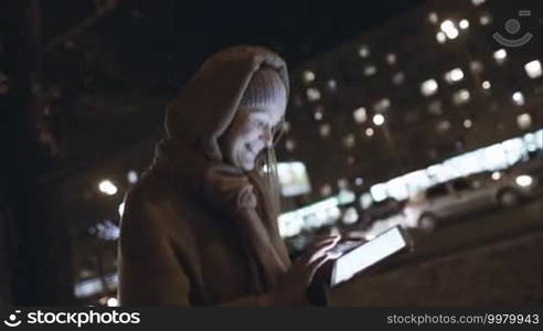 Steadicam shot of a young smiling woman typing on a tablet PC, she is walking quickly in the city on a cold autumn night