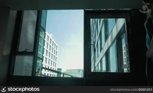 Steadicam shot of a young woman walking with a roll-on bag in the airport or hotel. She is passing by the windows with a view of a modern building