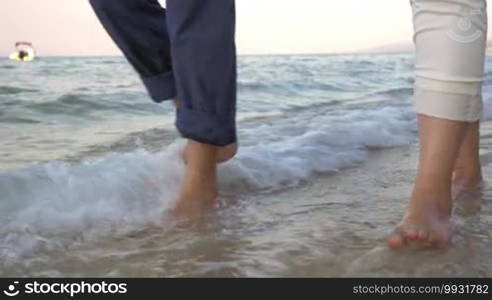 Steadicam shot of bare feet of two people walking along the sea in incoming waves.