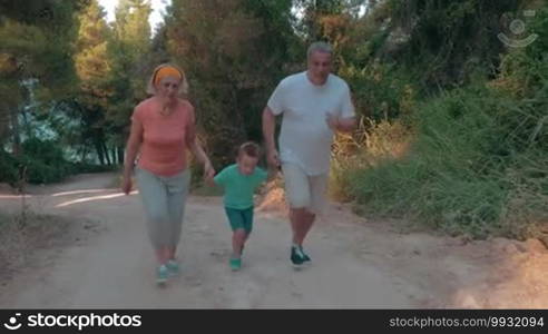 Steadicam shot of grandmother, grandfather, and little grandson running hand in hand up the road in the forest. Evening training together