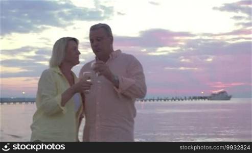 Steadicam shot of loving mature couple enjoying a romantic evening on the seashore. They are walking along the coast and having wine