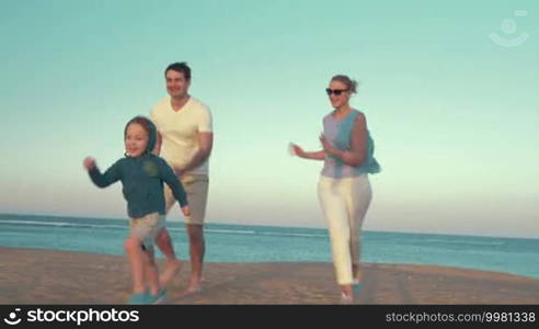 Steadicam shot of mother and father running a race with son on the beach. Boy running up to broken buoy and kicking it, parents applauding the little winner