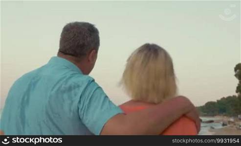Steadicam shot of senior couple enjoying an evening walk on the beach, man embracing woman as they go along the sea