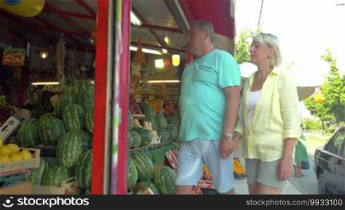 Steadicam shot of senior family couple selecting watermelon on street market. People squeezing it and rapping to make sure they choose ripe one