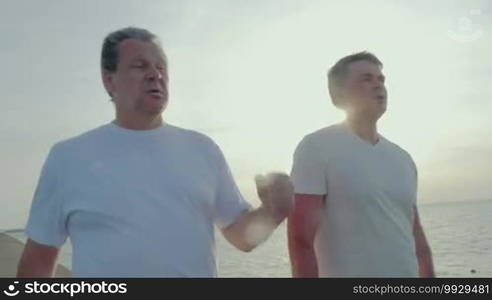 Steadicam shot of tired father and son walking after morning exercises, son is drinking water from plastic bottle.