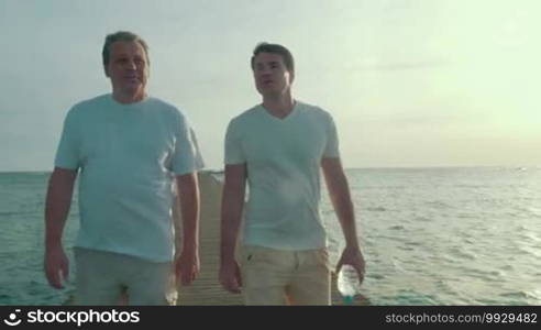 Steadicam shot of two friends or father and adult son having conversation while walking along the wooden pier in the sea