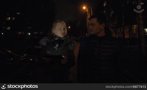 Steadicam shot of young couple having a walk outdoors in winter evening. They are talking, discussing something, and the woman is using a digital tablet on the way