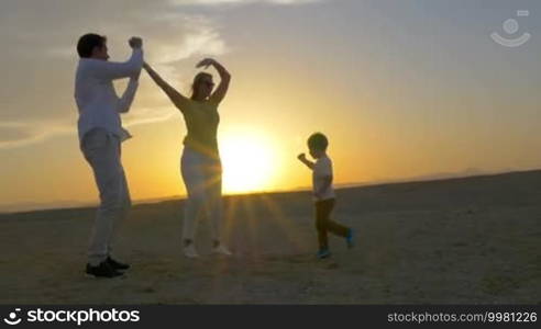 Steadicam shot of young happy parents and little son having fun while dancing on the beach at sunset. Bright evening sun shining in background. Summer vacation with family