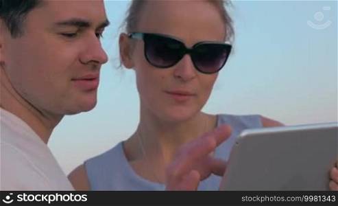 Steadicam shot of young man and woman outdoors with a tablet. They are enjoying the sunset reflecting in the woman's sunglasses