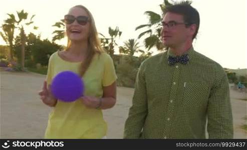 Steadicam shot of young woman walking with hipster style man on tropical resort at sunset. Happy girl playing with violet balloon while having a vivid talk with a friend