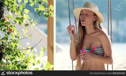 Stunning blonde woman on summer vacation sitting on a swing on the beach, smiling and having fun blowing out bubbles.