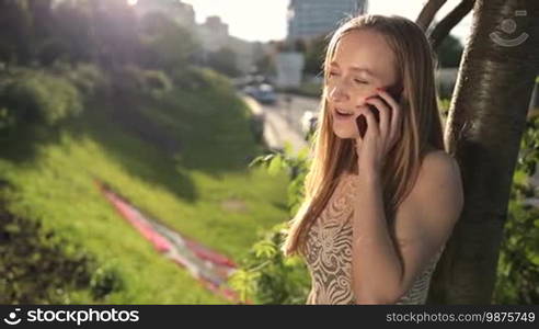 Stunning woman with amazing long blonde hair communicating on smartphone while leaning on the tree in the park. Charming lady talking on mobile phone outdoors in the flow of warm sunset. Slow motion. Steadicam stabilized shot.