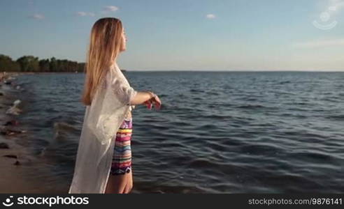 Stunning young blond woman taking deep breath enjoying fresh air in rays of beautiful sun on the beach during summer holidays. Delightful girl relaxing at seaside with arms raised outstretched up.