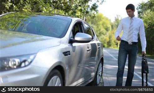 Stylish business executive in formal wear holding bag, unlocking car alarm system with remote control and getting into parked luxury vehicle over beautiful summer landscape background. Businessman going on business road trip by auto.