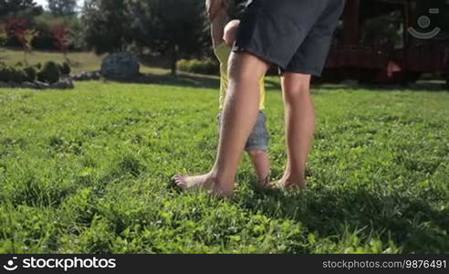 Sweet infant baby boy taking first steps barefoot with the help of affectionate father on green park lawn. Back view. Low view. Cute toddler child with caring dad learning to walk on grassy field in summer park. Slow motion. Steadicam stabilized shot