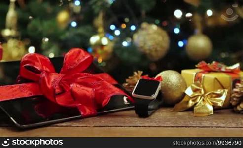 Tablet PC, smartphone, and smartwatch with gifts and decorations in front of a Christmas tree with lights on a wooden table.