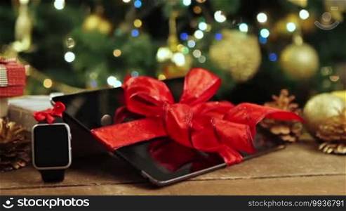 Tablet PC, smartphone, and smartwatch with gifts and decorations in front of a Christmas tree with lights on a wooden table.