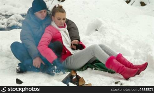 Teenage Couple Enjoying Winter Holidays by Bonfire in Snowy Forest