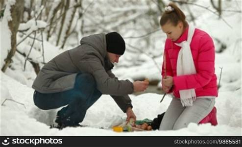 Teenage couple sitting by bonfire and roasting sausages in winter forest