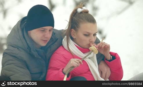 Teenage couple sitting by bonfire and roasting sausages in winter forest