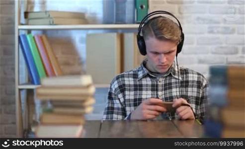 Teenage student with headphones taking online learning course on his smartphone at home. Attractive young man browsing cellphone and listening to music with headphones while having a break from studying.