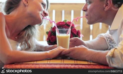 The bride and groom drink orange juice from one glass at a cafe