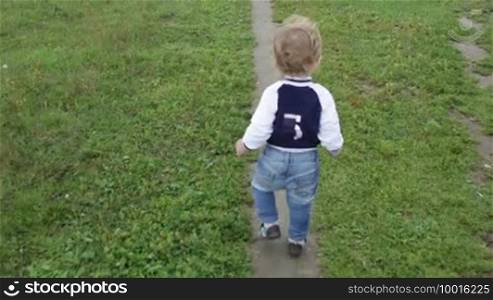 The one-year-old cute fair-haired boy in a blue jumper is taking his first steps. Shot from the back with camera stabilization equipment.