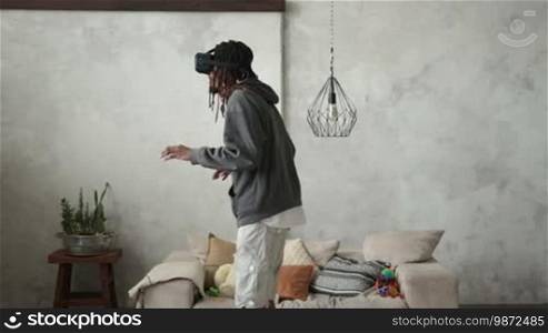 The picture in virtual reality glasses seems so real to young African man that he carefully minds his step. Full length of amazed African American hipster with dreadlocks with VR headset playing video games. Headset VR device playing with smartphone.