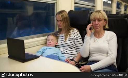 Three passengers in the train in the evening. Mother and son watching a movie on the computer, grandmother having a phone talk