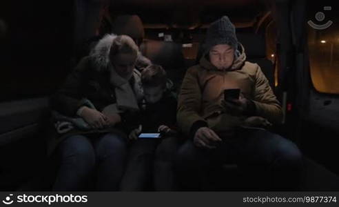 Three people having a ride in a minibus at night. Mother and son playing on a mobile while a male passenger surfs the internet on a cell phone and looks out the windows