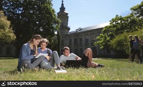 Three teenage hipster friends studying on campus lawn, using laptop and digital tablet PC while sitting on green grass outside university building. College students learning together with the help of modern technology devices. Dolly shot.